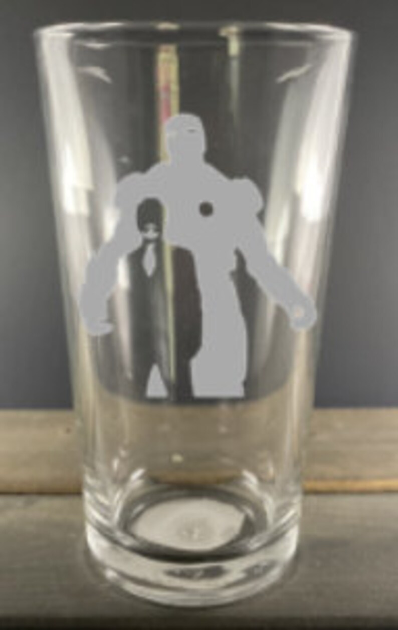 Puede incluir: Un vaso de pinta de vidrio transparente con una silueta blanca de Iron Man y otra figura de pie detr&aacute;s de &eacute;l.
