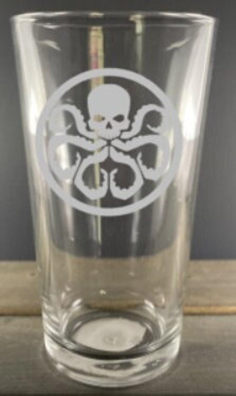 May include: A clear glass pint glass with a white etched design of a skull with tentacles in a circle. The design is reminiscent of the logo for the fictional organization HYDRA from the Marvel Cinematic Universe.