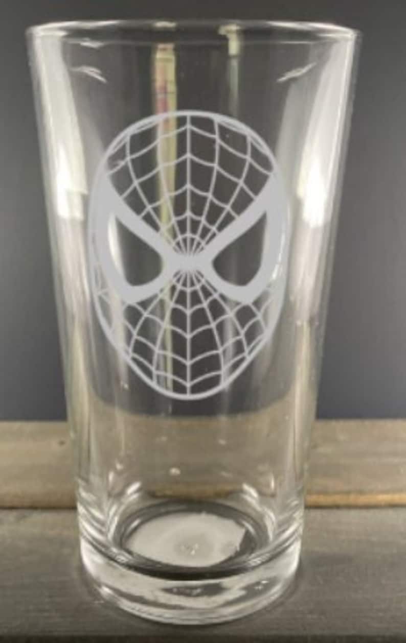 Puede incluir: Un vaso de pinta de vidrio transparente con un dise&ntilde;o grabado en blanco de la m&aacute;scara de Spider-Man.