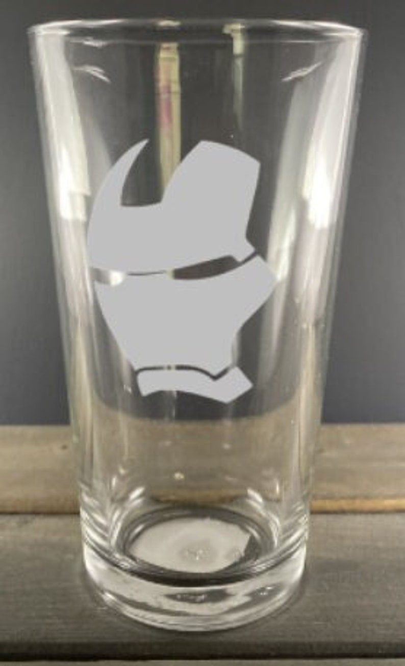 Puede incluir: Un vaso de pinta de vidrio transparente con una silueta blanca de la m&aacute;scara de Iron Man grabada en la parte delantera.