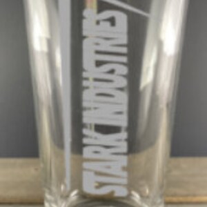 Puede incluir: Un vaso de pinta de vidrio transparente con el texto "Stark Industries" grabado en el lateral en blanco, con una fuente inclinada.