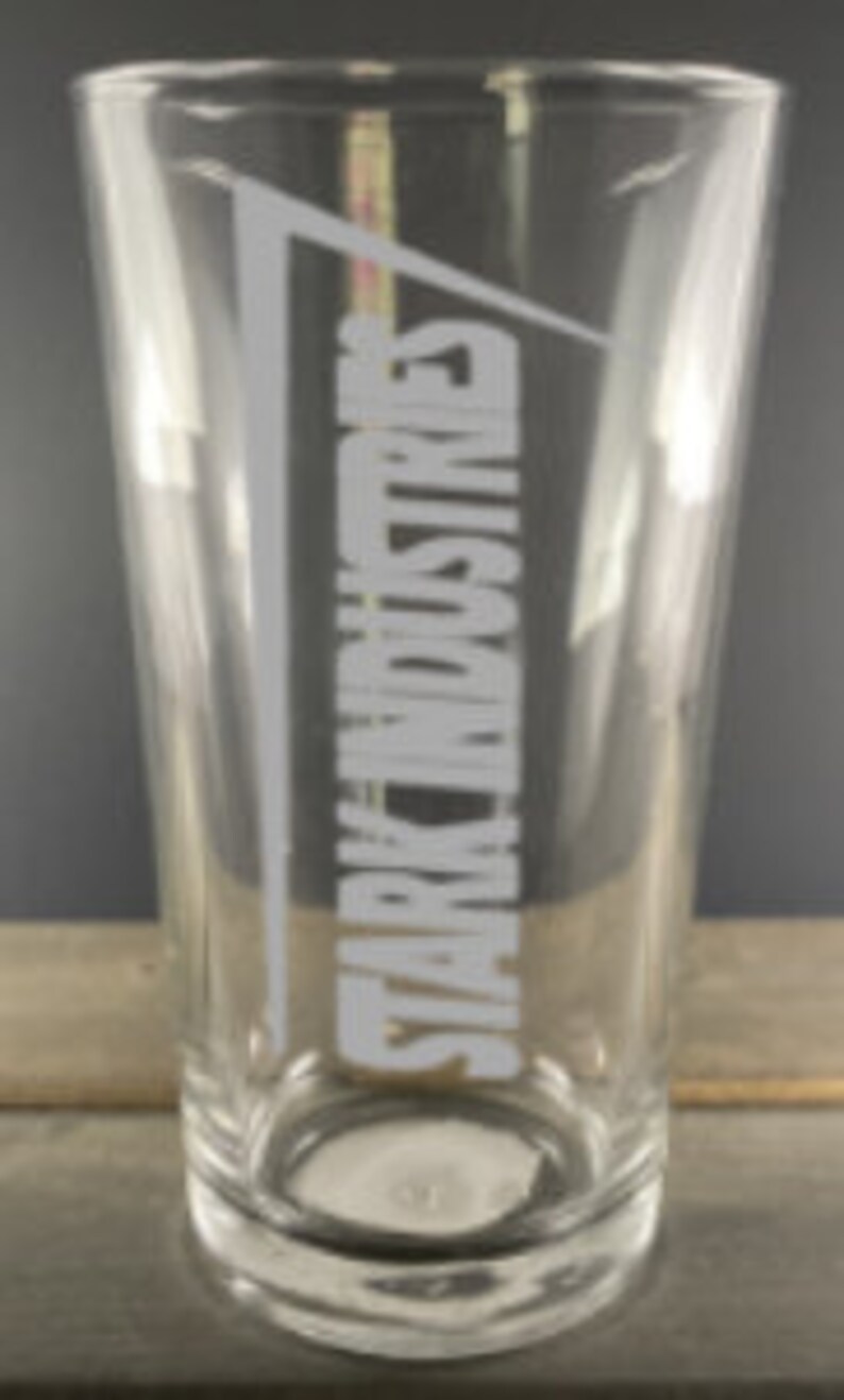 Puede incluir: Un vaso de pinta de vidrio transparente con el texto "Stark Industries" grabado en el lateral en blanco, con una fuente inclinada.