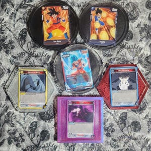 May include: A selection of anime trading cards, each depicting a different character, presented in various holders. The cards feature characters such as Son Goku and Vegeta. The holders are in black, clear, red, purple, and grey.