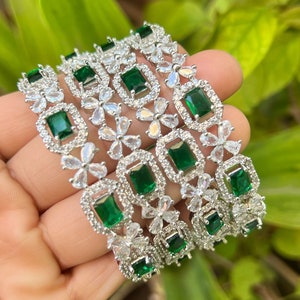 Könnte beinhalten: Ein Set aus fünf silbernen Armreifen mit smaragdgrünen und klaren Zirkonia-Steinen. Die Armreifen sind mit einem floralen Design verziert.