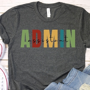 May include: A dark grey t-shirt with the text "ADMIN assistant" in colourful block letters. The letters are in shades of green, red, yellow, blue, and green.