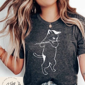 May include: A gray t-shirt with a white outline of a cat playing a flute. The text "Shop Salon" is on a circular tag with a floral design. The text "Hand Made With Love" is on the tag.