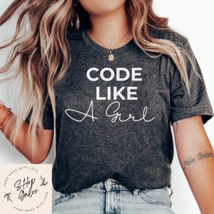 May include: Dark gray t-shirt with the text "CODE LIKE A Girl" in white. The shirt has short sleeves and is made of a soft material. The image also includes a small logo that says "Shop Galon".