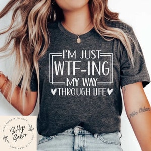 May include: Gray t-shirt with white text that reads "I'm just WTF-ING my way through life" with a heart on each side of the text. The shirt is being worn by a person with long blonde hair.