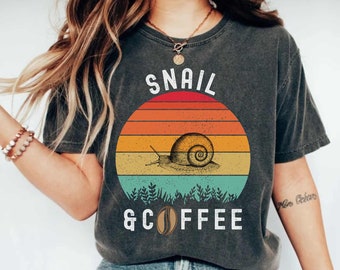 May include: A dark grey t-shirt with a vintage-style graphic of a snail and a coffee bean. The graphic features a sunset with orange, yellow, and blue stripes. The text "Snail & Coffee" is printed below the graphic.