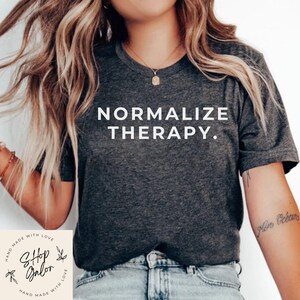 May include: Dark gray t-shirt with the words "NORMALIZE THERAPY." in white block letters. The shirt is a crew neck style and appears to be made of a soft material. The image also includes a small logo and the words "Shop Galon".