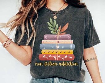 May include: A dark gray t-shirt with a stack of colorful books tied with twine and a sprig of leaves. The text "non fiction addiction" is printed below the books.