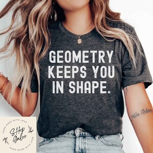 May include: A dark grey t-shirt with the text "GEOMETRY KEEPS YOU IN SHAPE." printed in white. The shirt is being worn by a person with long blonde hair.