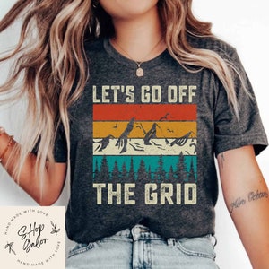 May include: A dark grey t-shirt with a vintage-style graphic design featuring mountains, trees, and the text "LET'S GO OFF THE GRID".