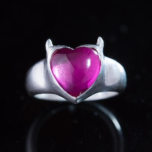 May include: A silver ring with a heart-shaped pink gemstone and two small horns on the top.