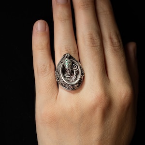 Op de afbeelding: Een zilveren ring met een gedetailleerd ontwerp met een skelet dat een bisschopsmijter draagt en een staf vasthoudt. De ogen van het skelet zijn bezet met groene edelstenen.