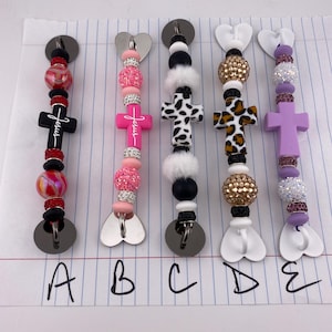 May include: A collection of five beaded pacifier clips with various designs. Each clip features a cross pendant, decorative beads, and a metal clip. The clips are in different colors, including pink, purple, and black, with the word "Jesus" on some.