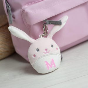 May include: A plush light pink rabbit keyring featuring long ears, embroidered facial features, and a white lower section displaying the capital letter M in pink. The keyring is attached to a silver metal ring and clip, hanging from a pink backpack.