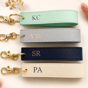 May include: Four personalised keychains in pastel shades: mint green, grey, navy blue, and cream. Each keychain has gold-coloured hardware and initials: KC, VW, SR, and PA. The keychains are arranged on a light-coloured surface.