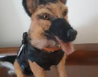 May include: A handcrafted German Shepherd dog figurine with a black vest and collar. The dog has brown and black fur, with its tongue sticking out. The figurine is made of felted wool.