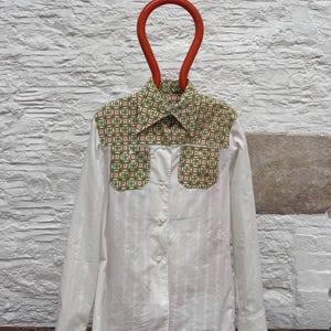 May include: White button-down shirt with a patterned collar and pocket details. The pattern features a repeating design of pink and green floral shapes. The shirt is hanging on an orange hanger.