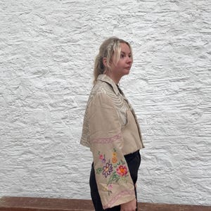 May include: A beige jacket with floral embroidery on the sleeves. The cropped jacket features a cream-coloured pattern on the upper section. The sleeves are decorated with colourful embroidered flowers and pink trim.