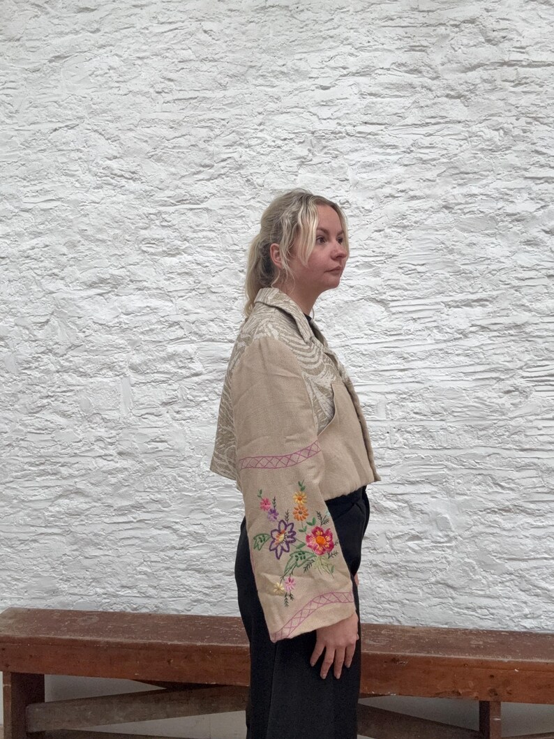 May include: A beige jacket with floral embroidery on the sleeves. The cropped jacket features a cream-coloured pattern on the upper section. The sleeves are decorated with colourful embroidered flowers and pink trim.