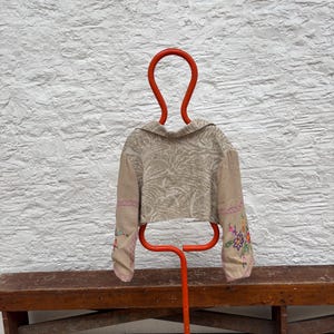 May include: A tan cropped jacket with floral embroidery on the sleeves is displayed on a bright orange metal hanger. The jacket has a collar and a patterned design. The hanger is in front of a white textured wall and a wooden bench.