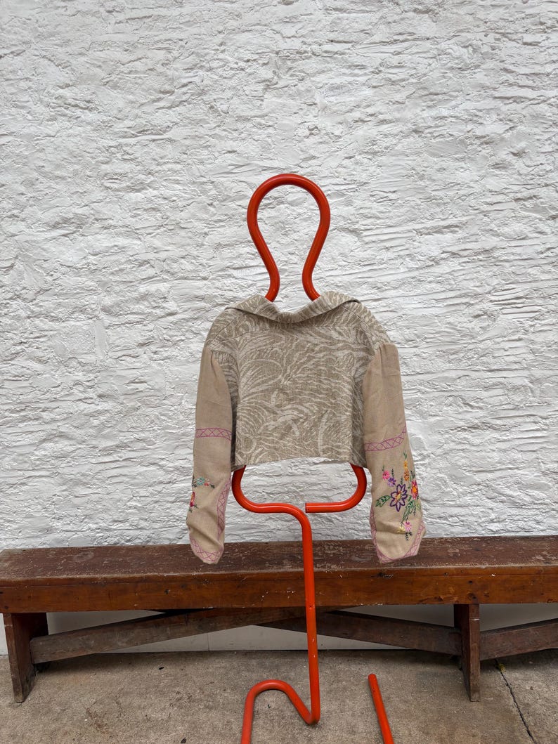 May include: A tan cropped jacket with floral embroidery on the sleeves is displayed on a bright orange metal hanger. The jacket has a collar and a patterned design. The hanger is in front of a white textured wall and a wooden bench.