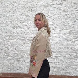 May include: Beige cropped jacket with floral embroidery on the sleeves. The jacket has a collared neckline and a patterned design on the upper back and shoulders. The sleeves are adorned with colorful embroidered flowers and pink stitching.