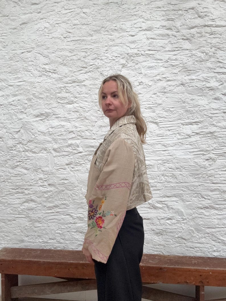 May include: Beige cropped jacket featuring floral embroidery on the sleeves. The jacket has a collared neckline and a patterned design on the upper back and shoulders. The sleeves are decorated with colourful embroidered flowers and pink stitching.