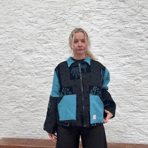 May include: A black jacket with teal blue panels and a patterned design. The jacket features a zip closure and two front pockets. The collar and sleeves are black, worn over black trousers.