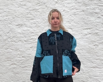 May include: A black jacket with teal blue panels and a patterned design. The jacket features a zip closure and two front pockets. The collar and sleeves are black, worn over black trousers.