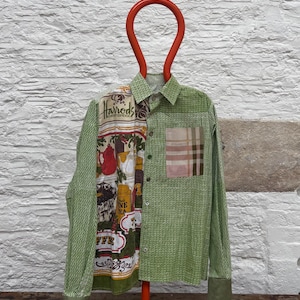 May include: A green shirt with a patchwork design. The left side has a print with the text "Harrods" and images of teapots. The right side is plain green with a plaid pocket. The shirt is hanging on a red hanger.
