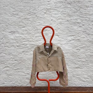 May include: A beige cropped jacket with floral embroidery and a button closure, hanging on a bright orange hanger. The sleeves feature pink stitching. The jacket is displayed against a textured white wall.