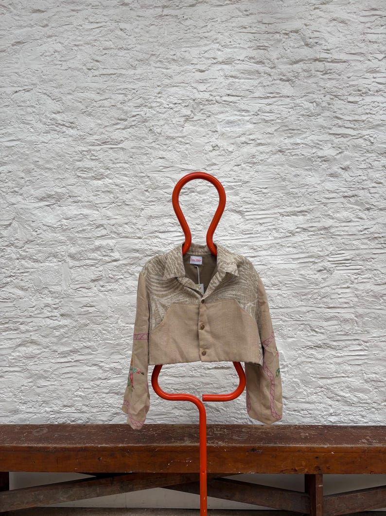 May include: A beige cropped jacket with floral embroidery and a button closure, hanging on a bright orange hanger. The sleeves feature pink stitching. The jacket is displayed against a textured white wall.