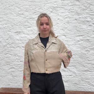 May include: A tan cropped jacket with floral embroidery on the sleeves and a patterned yoke. The jacket has a button-down front and a collared neckline. The model is wearing black trousers.