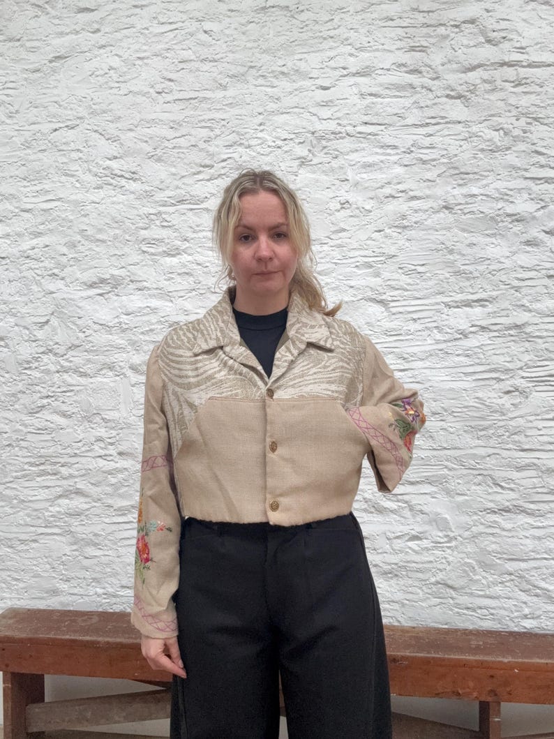 May include: A tan cropped jacket with floral embroidery on the sleeves and a patterned yoke. The jacket has a button-down front and a collared neckline. The model is wearing black trousers.