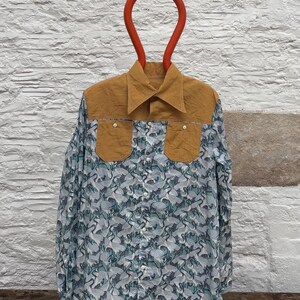 Può includere: Camicia button-down con carrè e tasche in velluto a coste color senape. Il corpo della camicia presenta un motivo di gru blu e bianco. La camicia è esposta su una gruccia rossa contro un muro di mattoni bianchi.