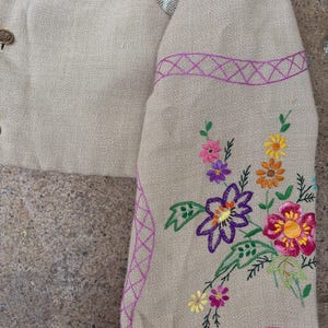 May include: Close-up of a beige jacket sleeve with colourful floral embroidery. The sleeve features a purple embroidered border and a variety of flowers in shades of pink, yellow, orange, and purple. The jacket has decorative buttons.