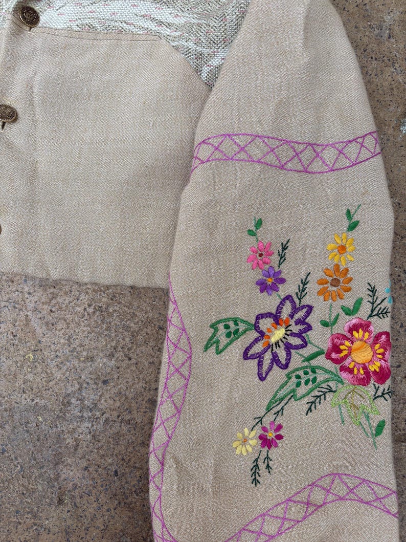 May include: Close-up of a beige jacket sleeve with colourful floral embroidery. The sleeve features a purple embroidered border and a variety of flowers in shades of pink, yellow, orange, and purple. The jacket has decorative buttons.