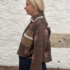 Può includere: Giacca corta marrone con ricami floreali rosa. La giacca presenta un colletto alto con finiture in pizzo bianco, una tasca marrone con finiture in pizzo bianco e maniche svasate. La giacca è indossata con pantaloni blu scuro.