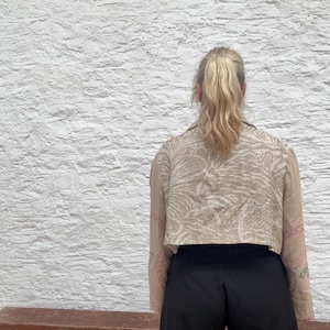 May include: A cropped beige jacket with a white and beige pattern and pink embroidery on the sleeves. The person is wearing black trousers. The background is a white textured wall.