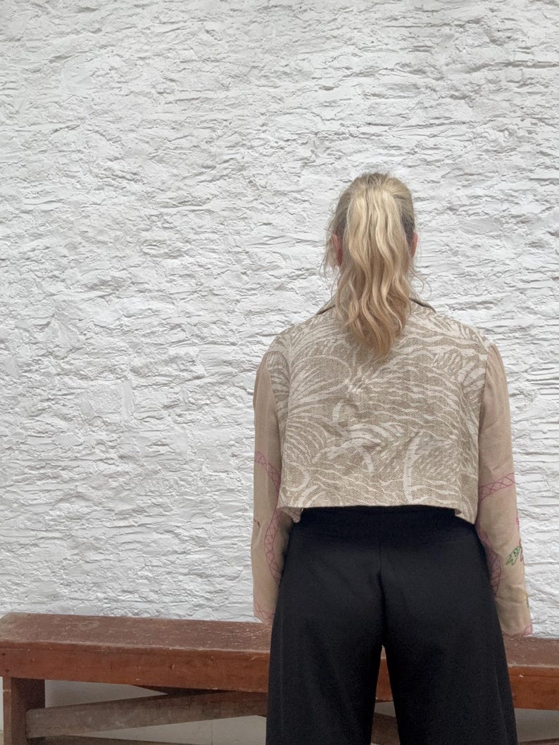May include: A cropped beige jacket with a white and beige pattern and pink embroidery on the sleeves. The person is wearing black trousers. The background is a white textured wall.