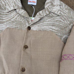 May include: A tan jacket with a patterned collar and upper chest. The jacket has three decorative buttons and a tag that reads "Deimos Atelier". The patterned section features shades of tan and white.