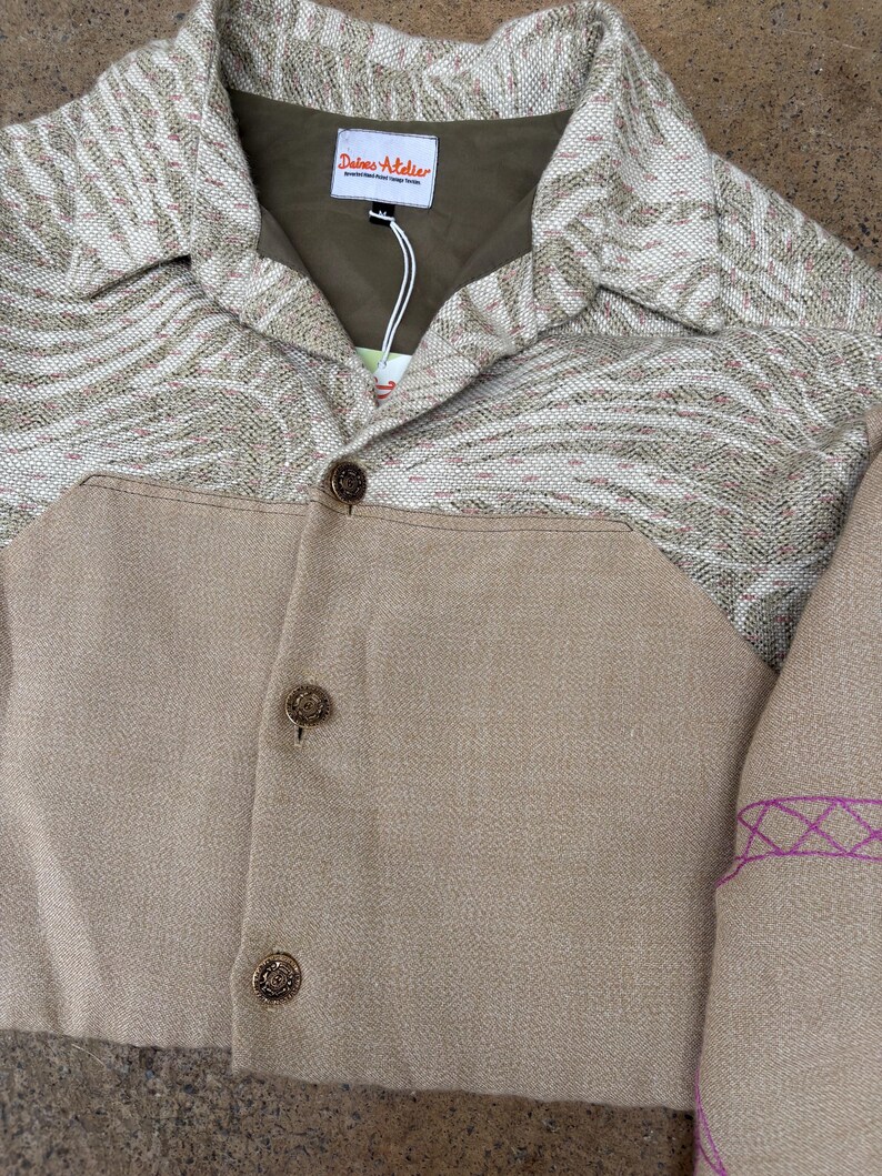 May include: A tan jacket with a patterned collar and upper chest. The jacket has three decorative buttons and a tag that reads "Deimos Atelier". The patterned section features shades of tan and white.