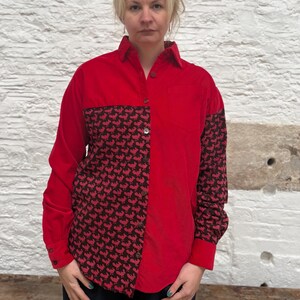 Può includere: Camicia rossa con un pannello a contrasto con motivo nero e rosso. La camicia a maniche lunghe ha un design con bottoni, un colletto classico e una tasca sul petto. Il motivo è un motivo ripetuto.