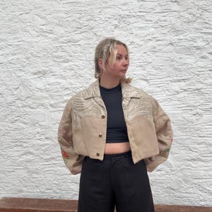 May include: Cropped beige jacket with floral embroidery on the sleeves and a zebra print pattern on the upper chest. The jacket has gold buttons and is worn with black trousers.