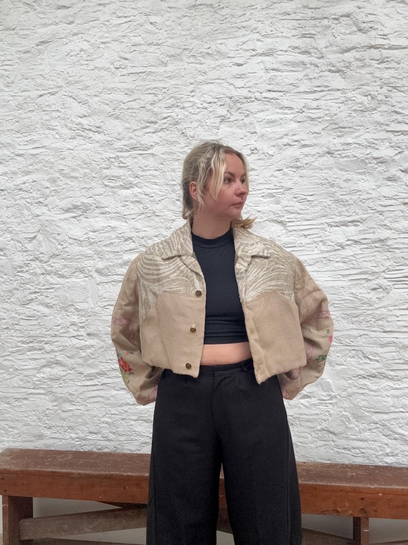 May include: Cropped beige jacket with floral embroidery on the sleeves and a zebra print pattern on the upper chest. The jacket has gold buttons and is worn with black trousers.