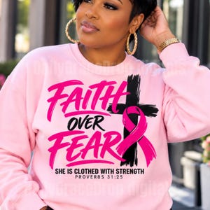 May include: A light pink long-sleeve sweatshirt featuring the black and pink text "FAITH OVER FEAR" with a black cross graphic wrapped in a pink ribbon. Below the text, it reads "SHE IS CLOTHED WITH STRENGTH PROVERBS 31:25" in black lettering.