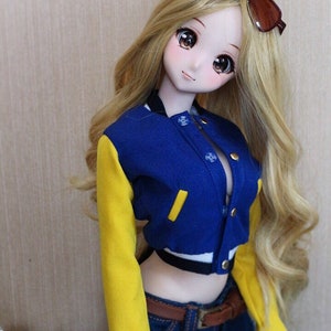 Pode incluir: Uma boneca com cabelo longo e loiro usando uma jaqueta de beisebol azul e amarela, jeans azul e um cinto marrom. A boneca está usando óculos de sol.
